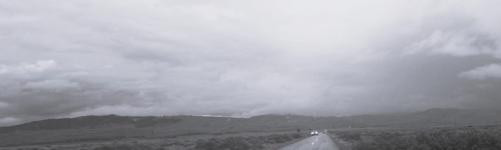 A lone automobile with headlights is driving toward the camera on a paved road amid clouds and fog.