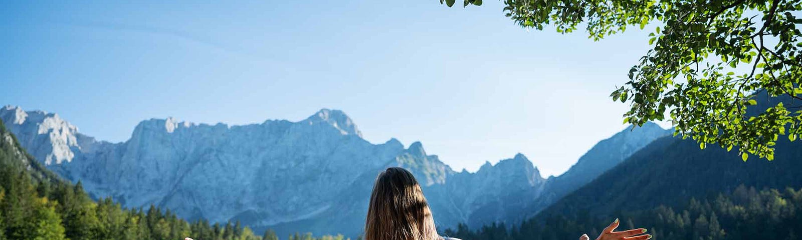 A woman stands with her arms wide open. She’s looking at a calming landscape of water and greenery.