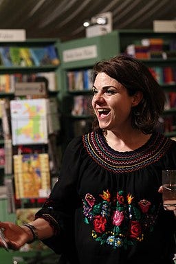 Caitlin Moran in three quarter profile with open grin, holding a glass of water in front of books, wearing black smock top