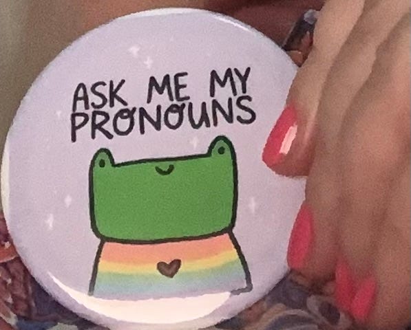 This a photo of a button owned by the author. The author is holding the button and you can see her fingers. The button has a light purple background with a smiling cartoon frog wearing a rainbow shirt with a small heart decoration. The text above the frog’s head says ASK ME MY PRONOUNS.