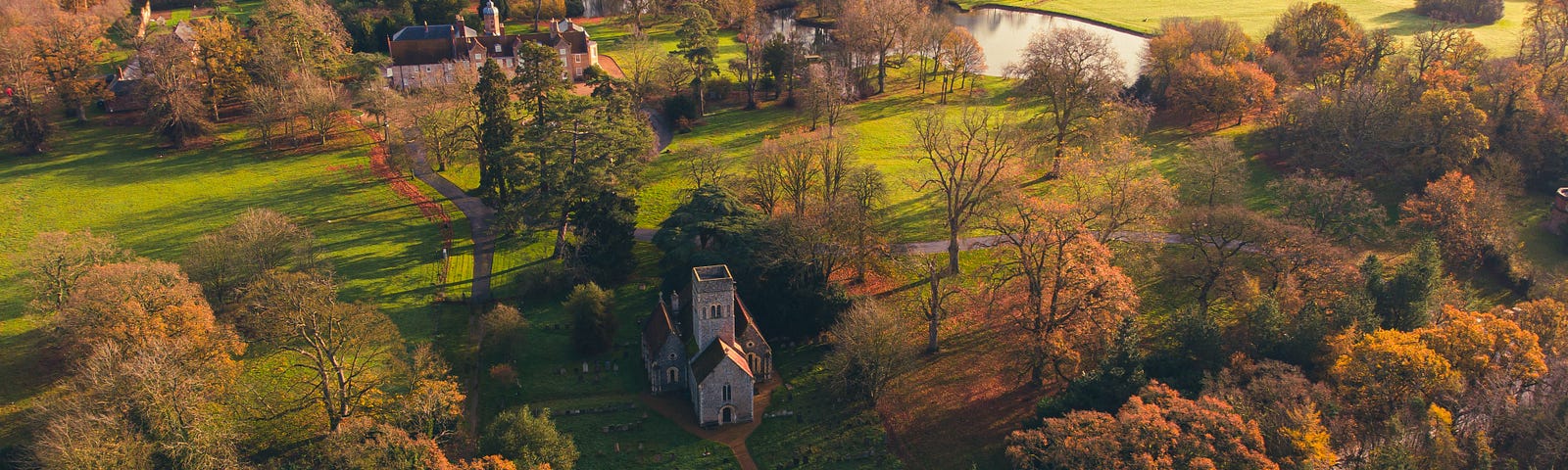 Aerial view of an English countryside with green grass, river, trees with autumn leaves, a road with two white vehivles and featuring St Mary’s Church, Gillingham, Norfolk
