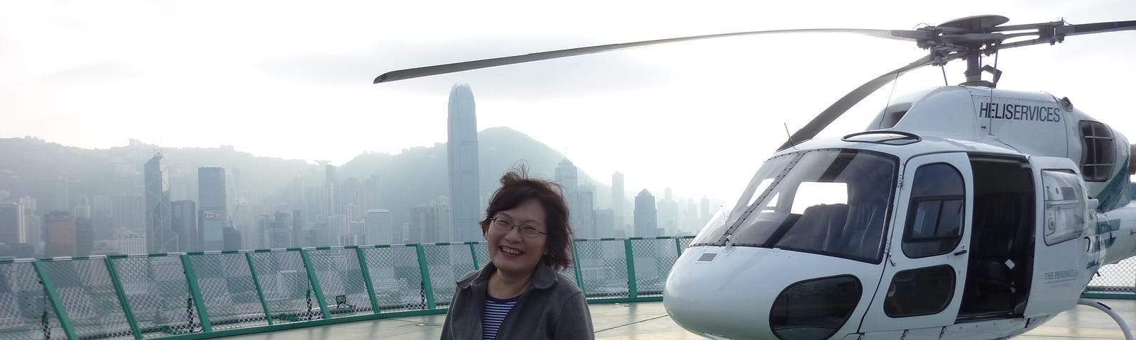 Author in front of a helicopter on top of a building in Hong Kong. The skyline is visible through a haze on the left side of the photo.