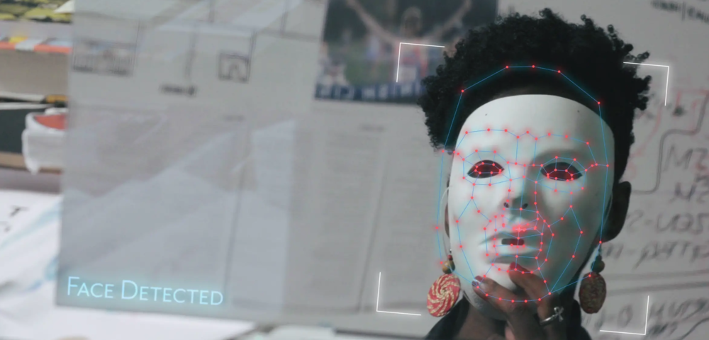 Photograph shows a screen that reflects a black woman holding a white mask over her face. The screen appears to show face detection technology at work, with lines mapping the mask, and text saying ‘Face detected’ in the bottom corner