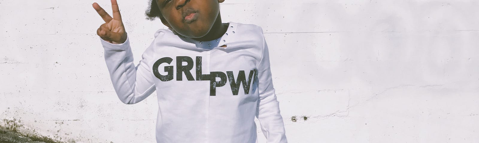 Kiana Bosman @capturedby_kiana portrait of young Black girl holding up a “peace sign” & wearing tee with words “GRL PWR.”