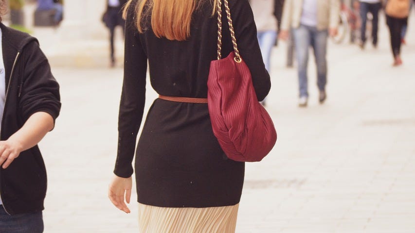 A woman wearing a stylish long, cream skirt and burgundy jumper, cinched at the waist with a thin, brown belt is walking away from us down a busy street.