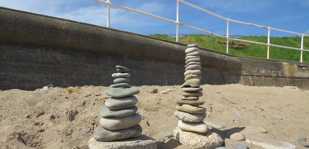 An “Inuksuk.” Two piles of stones about two feet high, on a seaside beach, with a sea wall, cliffs and blue sky in the background.