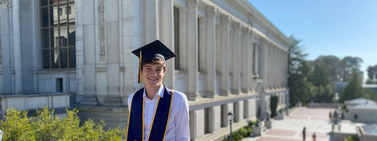 Young man poses with a stole and graduation cap in front of UC Berkeley’s Doe Library.