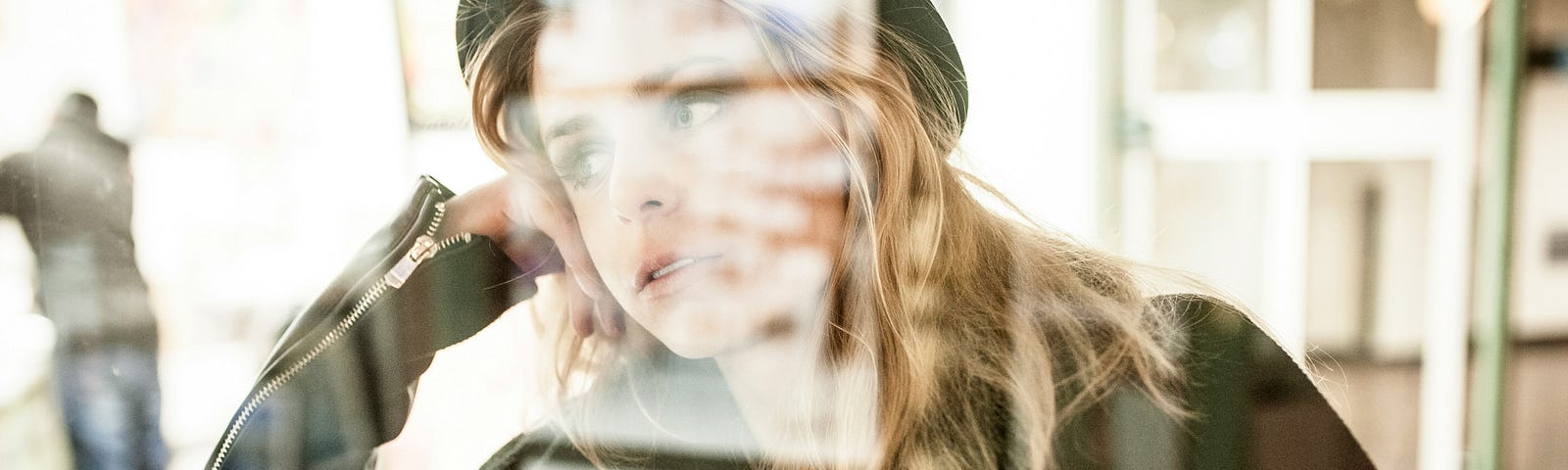A young blonde woman looks out a window. The outside world is reflected in the glass. She looks sad or contemplative. It’s a close-up shot.