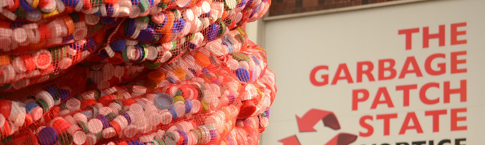 An image of a artwork — a spiral tower made of plastic bottle caps. In the background is a poster saying ‘The Garbage Patch State’ Vortice.