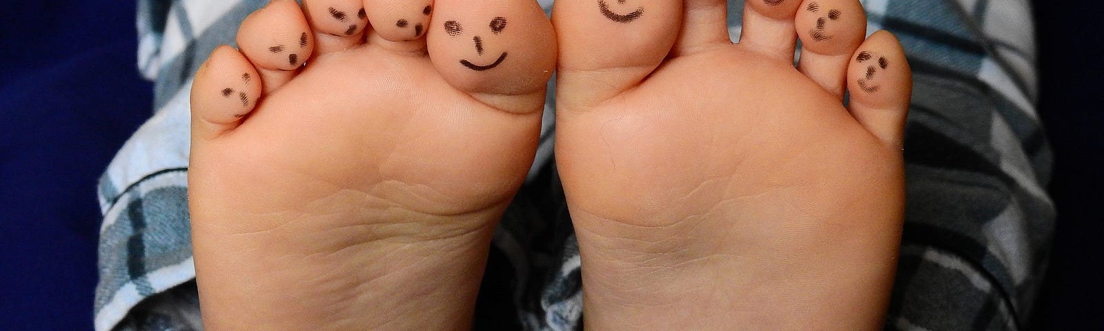 Photo of a child’s feet with black marker drawings of happy faces on each of their toes. Child is wearing black plaid pajamas