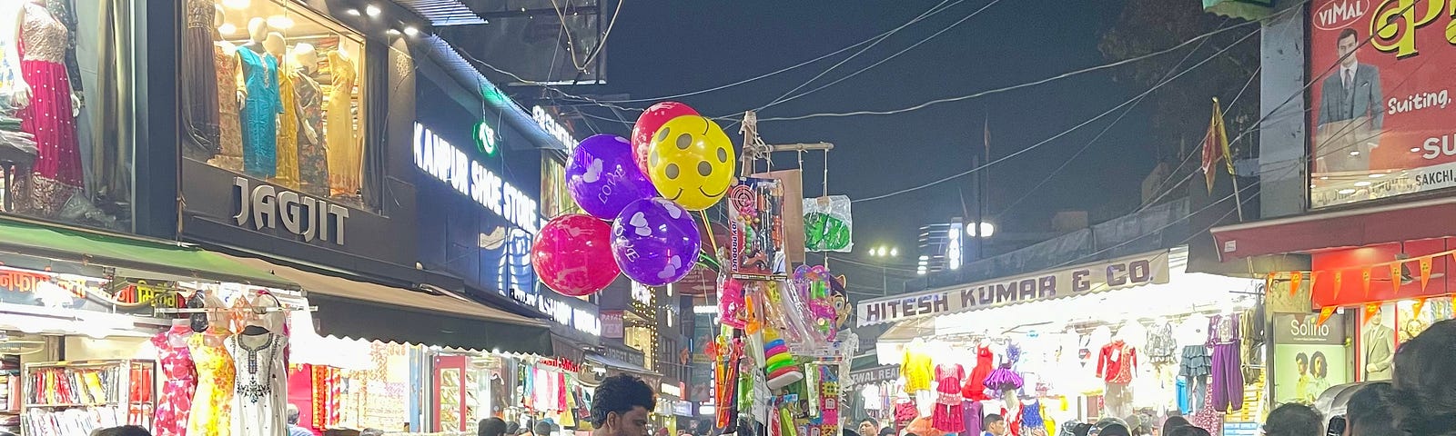 Motorcycles are parked in the center of this picture. Someone’s selling socks off one of the motorcycles. A toy seller sells balloons and toys. Clothes shops display their wares, There are plenty of people in this picture. Some people are walking around with their motorcycle helmets on.