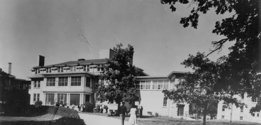 Black and white picture of thethree-story Assiniboia Residential School surrounded by trees, a road, a hedge, and a manicured lawn. Two people are walking towards the school, holding hands.