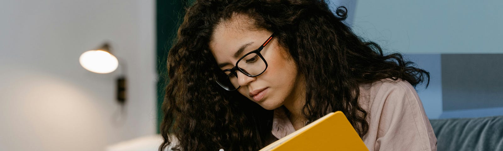 Woman with glasses and curly brown hair sitting on a couch writing in a yellow journal or notebook