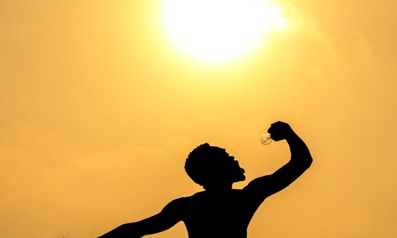 A silhouette of a man against a hazy sundown sky. One of his wrists is chained down. He is holding up a lightbulb in his other hand and looking at it with an open mouth, as if he’s going to eat it.