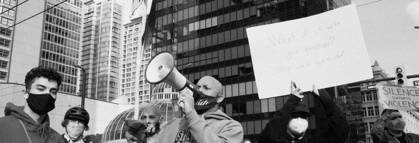 A black & white photo of a man in a crowd at the Black Lives Matter rally in Vancouver, BC, in May 2020. He’s wearing a grey hoodie and speaking into a megaphone. A masked person beside him is holding a sign that reads “What if it was your brother? What would you do?”