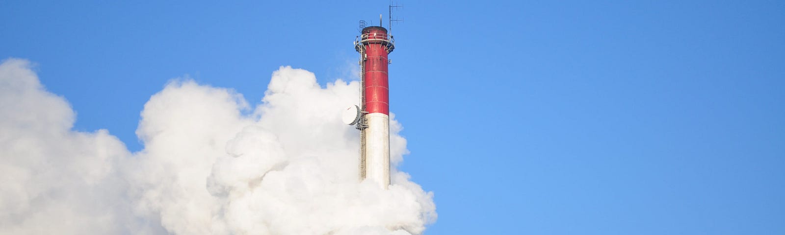 A close-up shot of a pair of red and white industrial chimneys against a blue, cloudless sky, with thick clouds of smoke billowing out of them.