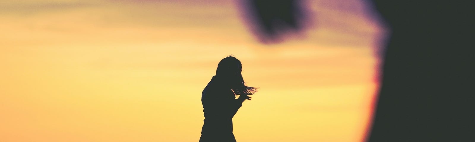 A apprehensive woman’s silhouette standing on a beach at sunset with an ominous figure in the forefront.