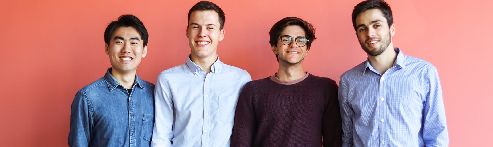 Four business casual dressed young men pose for a group photo in front of a pink wall.