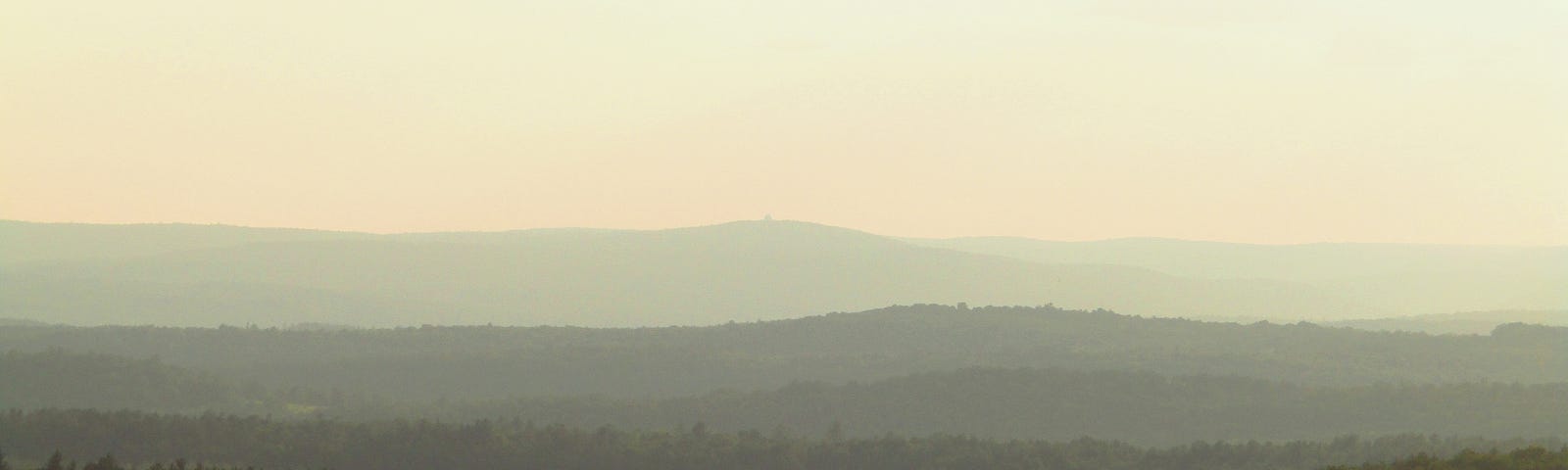 Layers of green forested hills fading into the distance, seemingly without end, beneath a pale, bright but hazy sky.