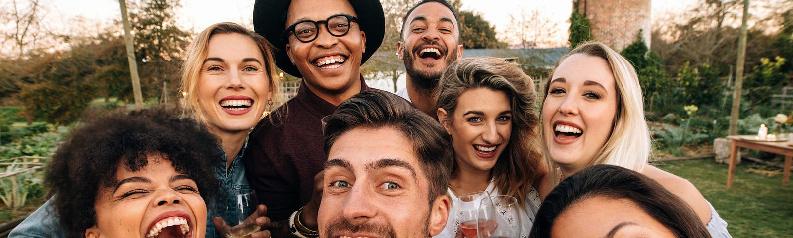 A group of 8 friends, a mix of races and genders, are gathered around a central figure who is holding a camera and taking a selfie. They are all smiling and laughing, clearing having a wonderful time together.