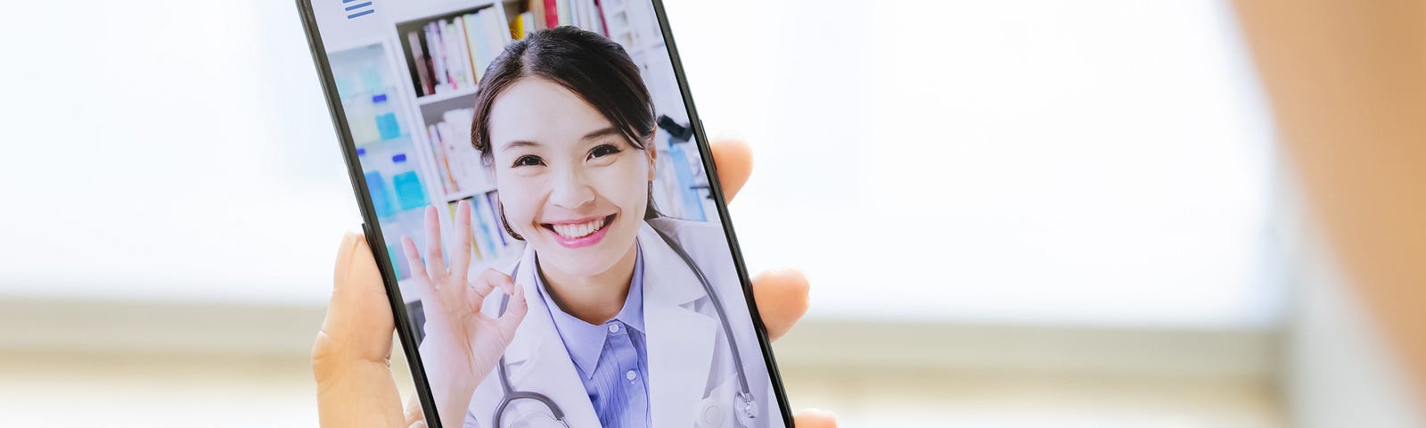 A woman holds a phone with a doctor on the screen for a telehealth appointment.