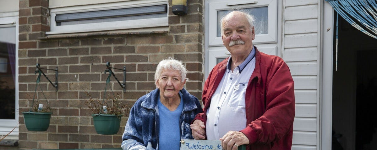 Photo of an older couple, Wilf and Carol as they stand outside a home. They hold hands and smile slightly at the camera.