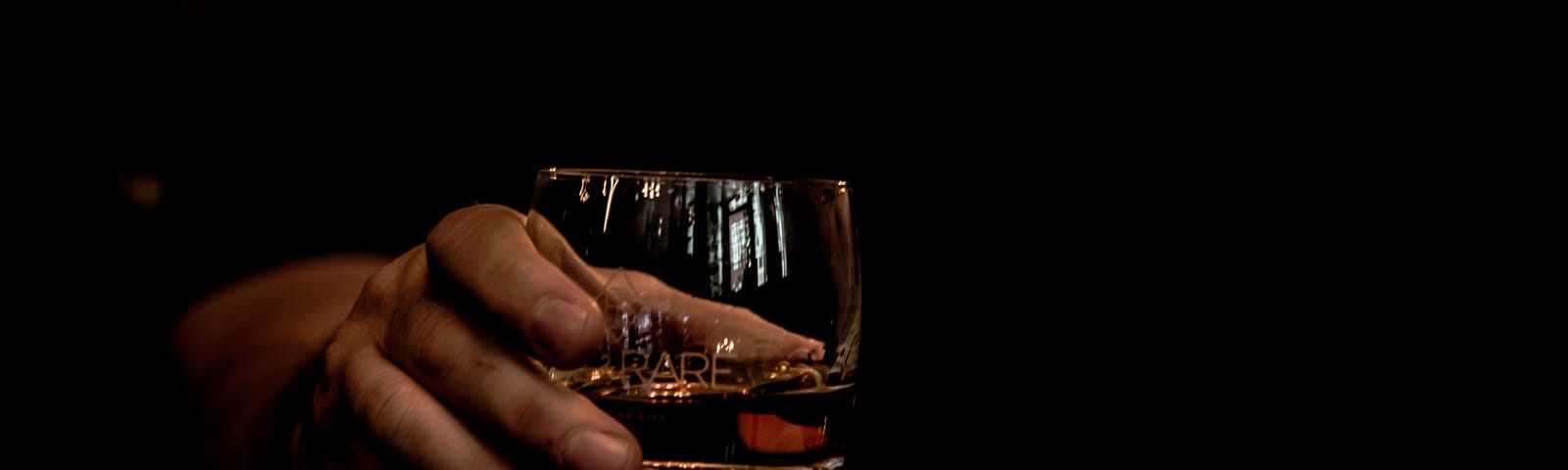 Dark picture with a hand holding a glass with whiskey in it.