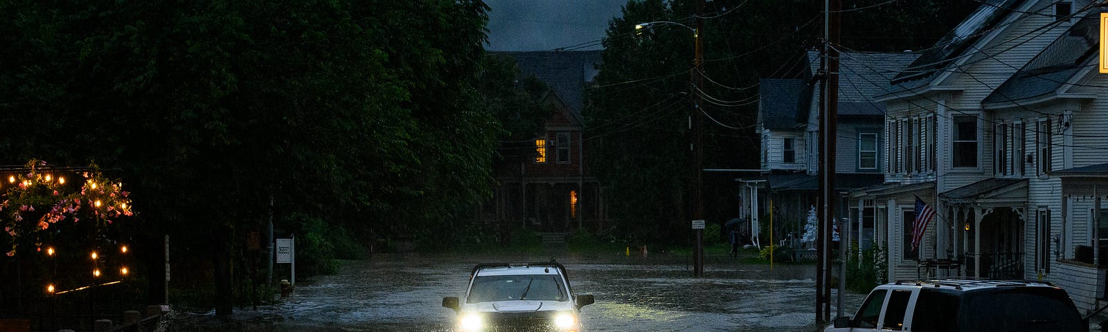 On a dark village street in the rain, headlights of an SUV shine barely above road level as the vehicle plunges through a flood