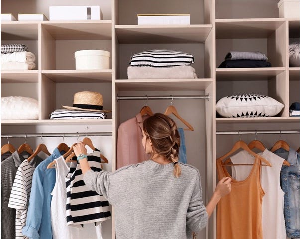 A woman stands facing a shelving unit on the back wall. At her head height, there is a bar with clothes hanging off. The clothes are light-coloured singlets, t-shirts, and jackets. Above this hanger are smaller box cabinets with white-coloured pillows, towels, and hats. The woman is reaching for a singlet on a hanger.