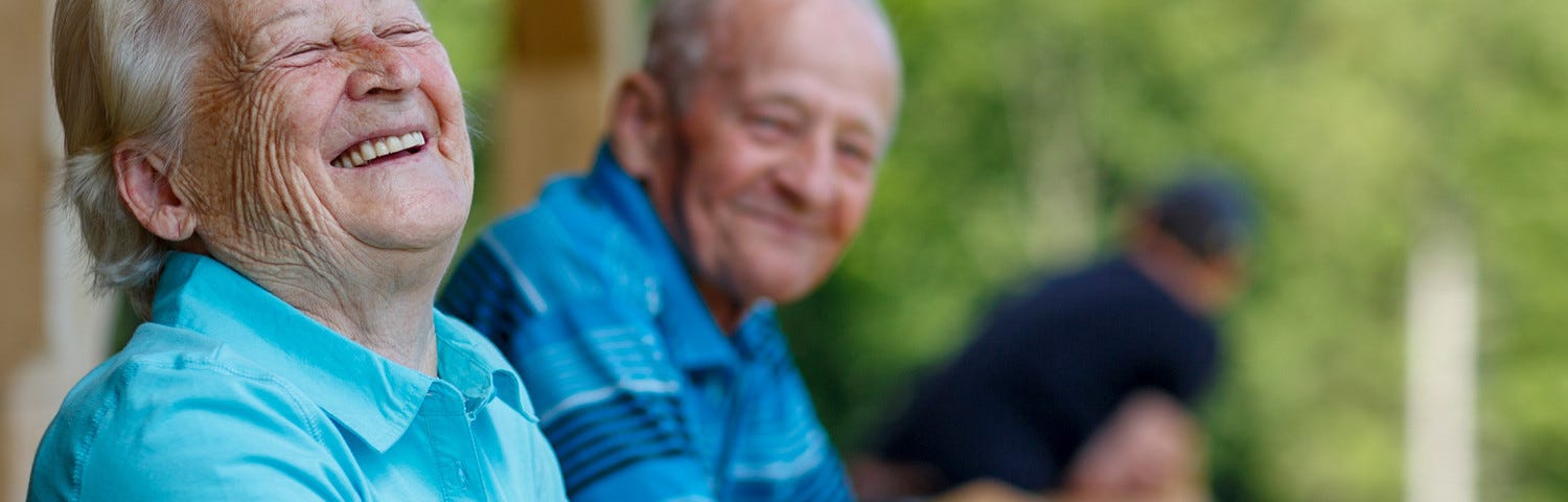 An older couple stand on a wooden balcony surrounded by trees. He is smiling and she has her head thrown back in laughter.