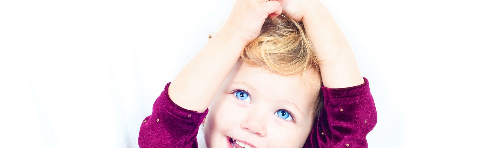 blonde toddler girl with big blue eyes and smile with arms on her head