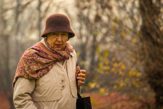 An older woman in a forest, wearing a fall coat, scarf, and wool hat.