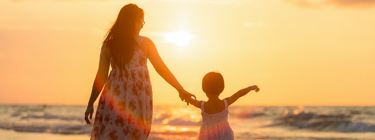 An adult and child walk hand in hand in the shallows of the ocean, toward the sunset.