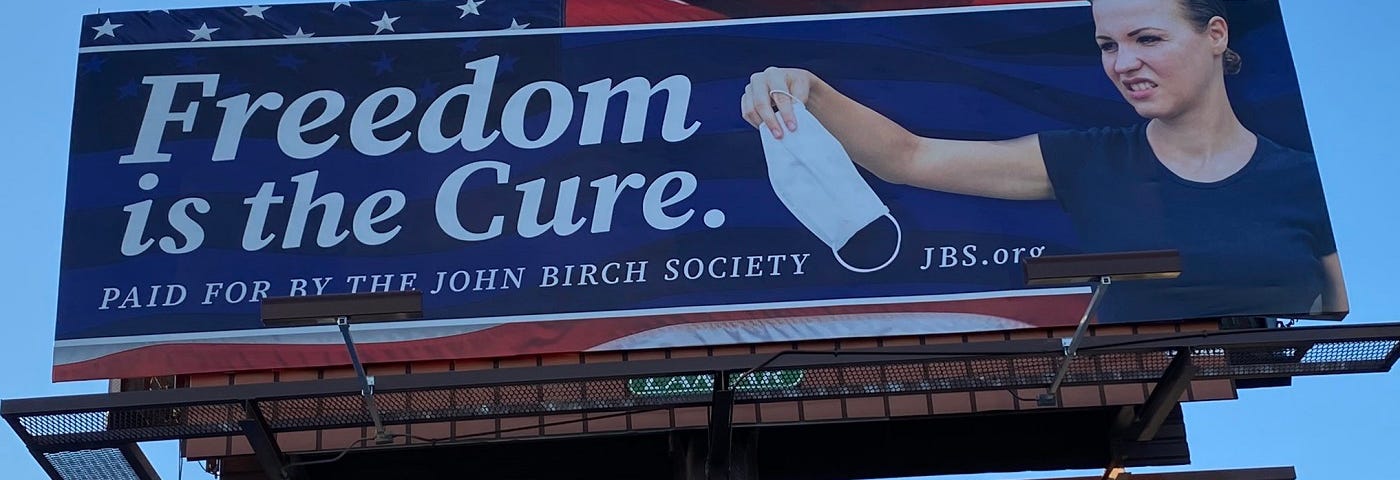 Photo of a highway billboard sponsored by the John Birch Society with a woman sneering and holding a face mask away from her. The words “Freedom is the Cure.