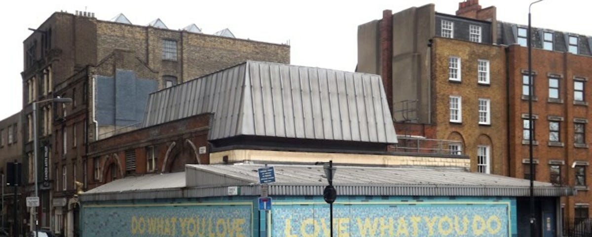 Two mosaic walls meeting at the corner of a city street. Both are pale blue with yellow lettering. One reads, “Do what you love,” and the other, “Love what you do.”