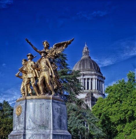 The Winged Victory Monument consisting of a bronze sculpture on a granite base is shown in front of the Washington State Legislative Building. Green foliage surrounds the scene.