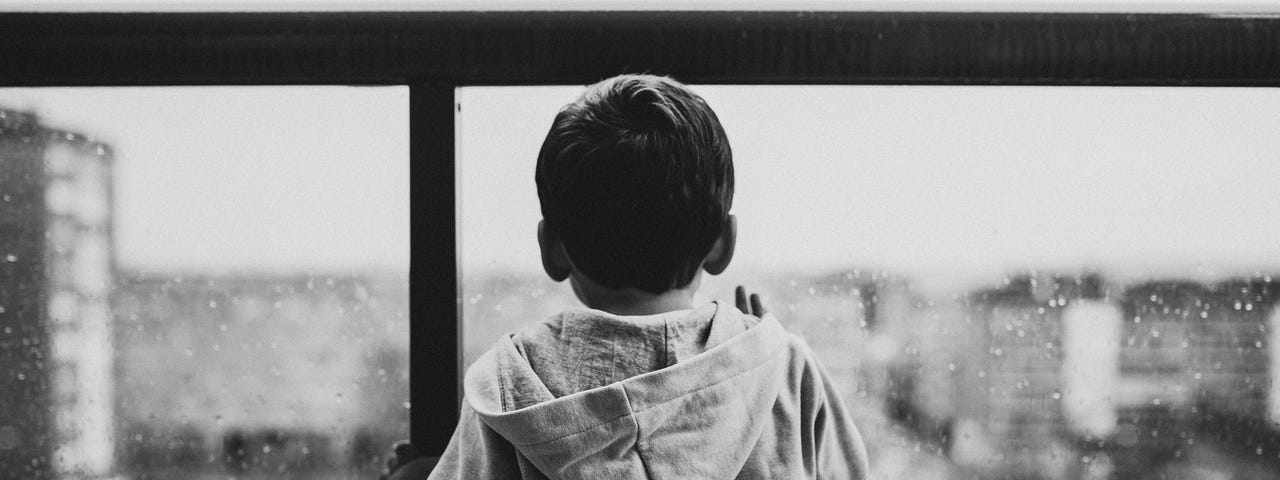 Black and white photo of boy wearing a hoodie while looking through a window on a rainy day in the city