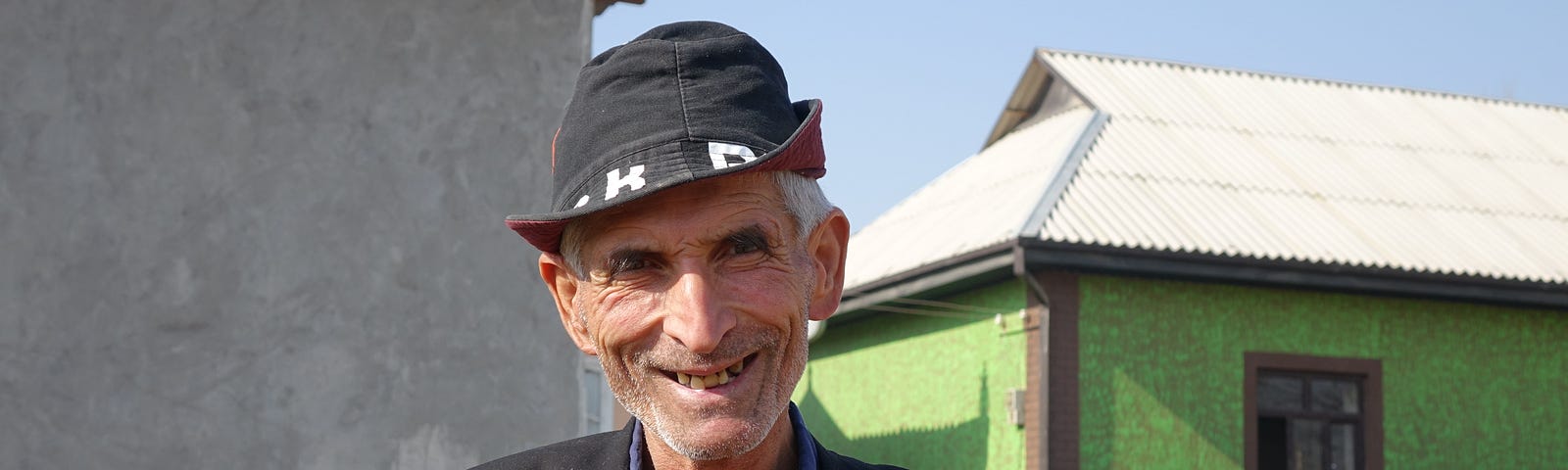 A older man with a wide smile and wearing a sharp black hat poses for a photo in front of his home.