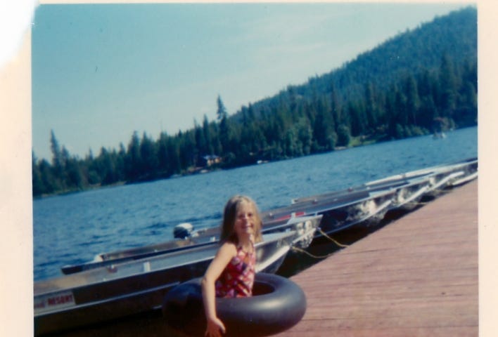 Young girl in a swimsuit inside an inner tube standing on a dock with boats moored at a lake.