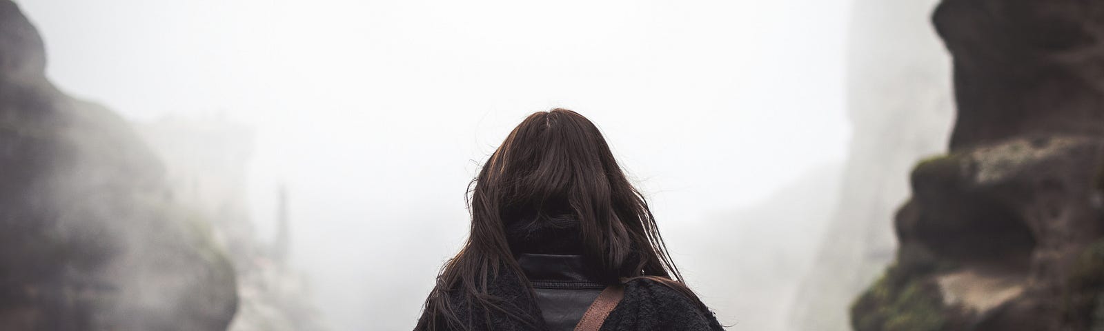 Woman with dark brown, shoulder-length hair standing between two rock walls with a leather satchel on her back heading into the fog.