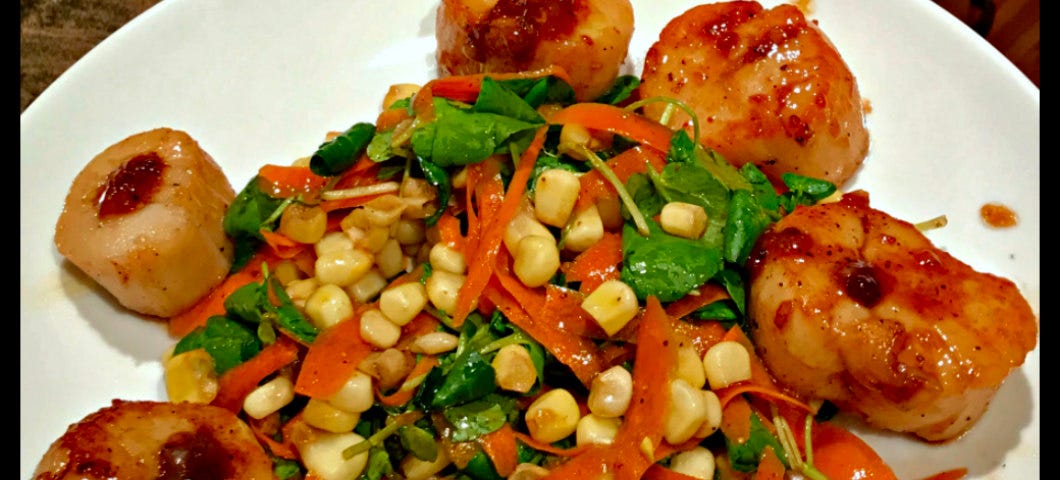 A colorful salad of corn, shredded carrots and greens surrounded by six caramelized scallops arranged on a white plate.