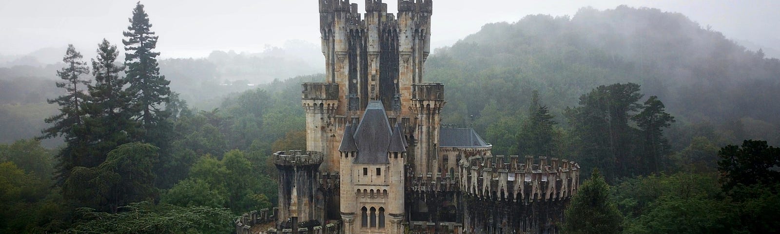 A small castle sits surrounded by woods. Misty hills int he background.