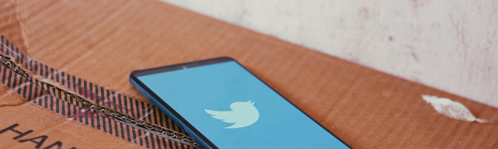 A phone with a blue screen and the twitter bird logo sits on a cardboard box that reads ‘handle with care’