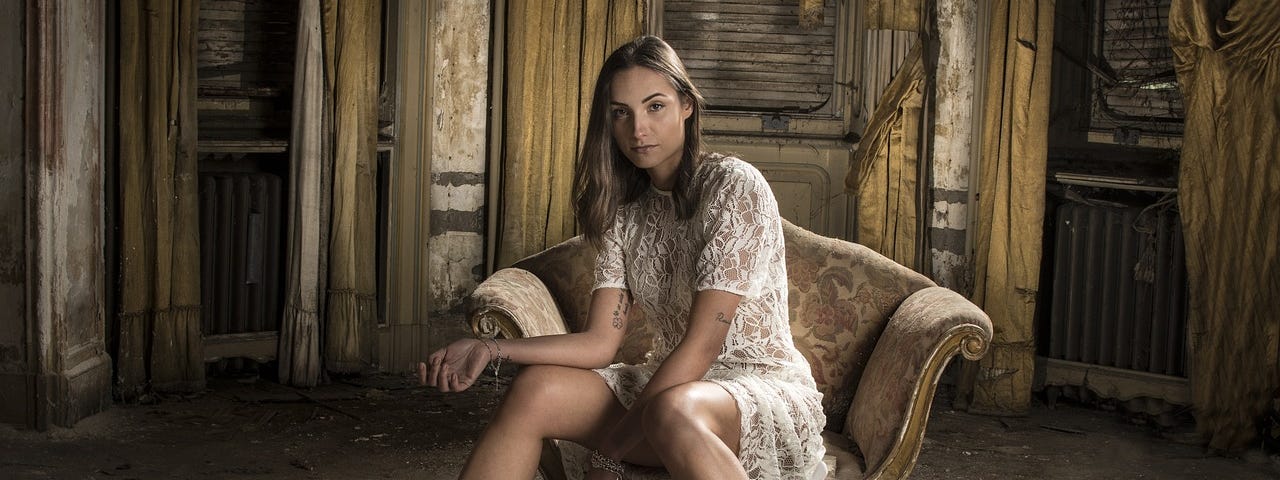 Image of a woman wearing a white lace dress and black high-heeled sandals, sitting on an old chair and surrounded by decayed walls inside a room.