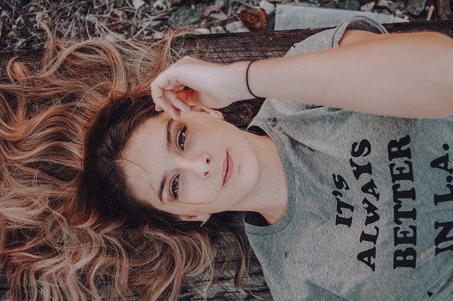 Young woman lies on her back, dark hair fanned out behind her head, wooden surface underneath, she wears grey t-shirt with logo, secret smile
