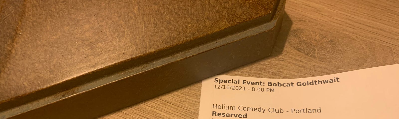Ticket stub for a comedy performance at Helium Comedy Club in Portland, OR, featuring Bobcat Goldthwait, December 16, 2021. the ticket is placed almost horizontal in the frame of the picture, the upper left corner of the stub is held down by the base of a lamp, and the surface beneath the ticket and lamp is wood grain particle board.