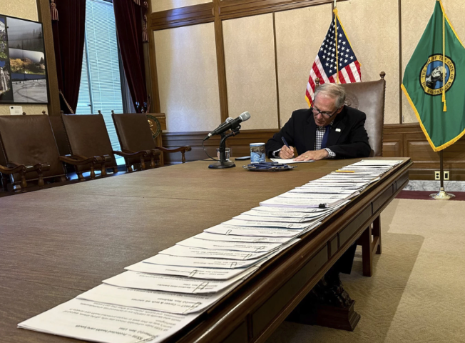 Governor Inslee signs a bill at the end of a long table. Lined up on one side of the tabletop is legislation that is waiting to be signed. A United States flag and a Washington State flag are displayed behind Inslee.
