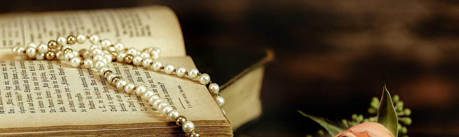 Color photo of pearl necklace draped over old book with pink rose via Pixabay