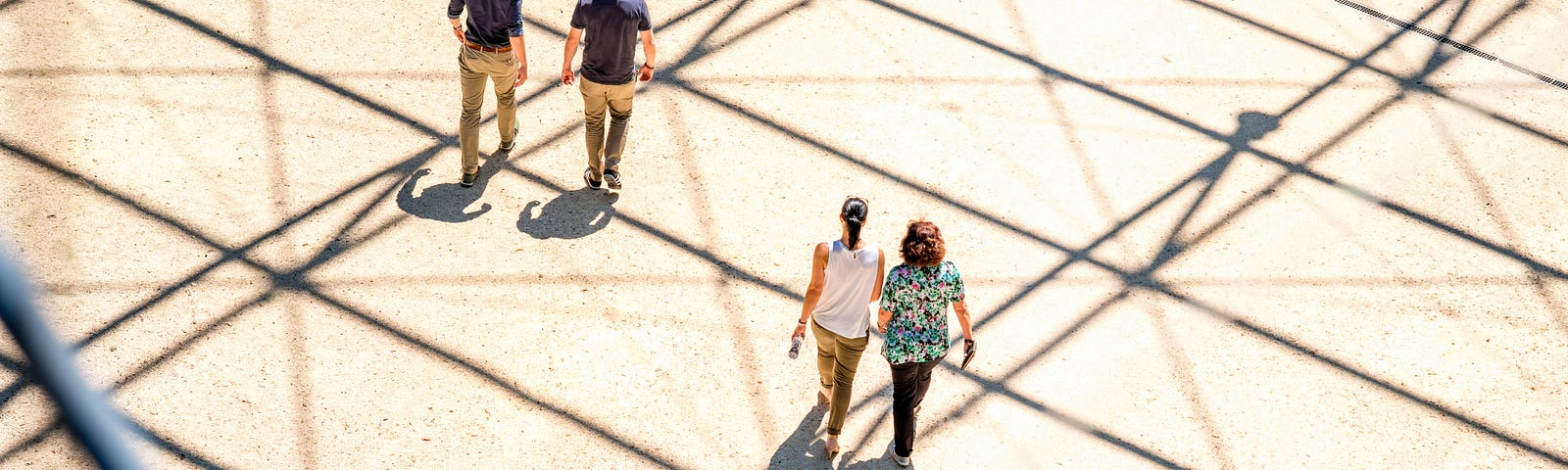 Two couples photographed from a distance, walking in an open area where long shadows cast from an unseen source create dark parallel lines on the ground.