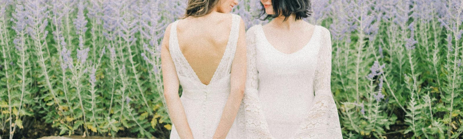 Two women stood outside in wedding dresses. Stood very close. One standing towards the camera, the other standing facing away from the camera. Looking at each other over their shoulders.